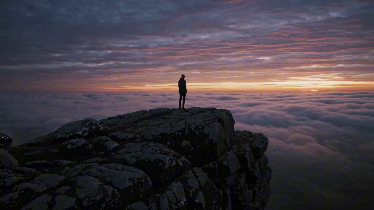 Silhouette of a person on a rocky cliff above clouds at sunset