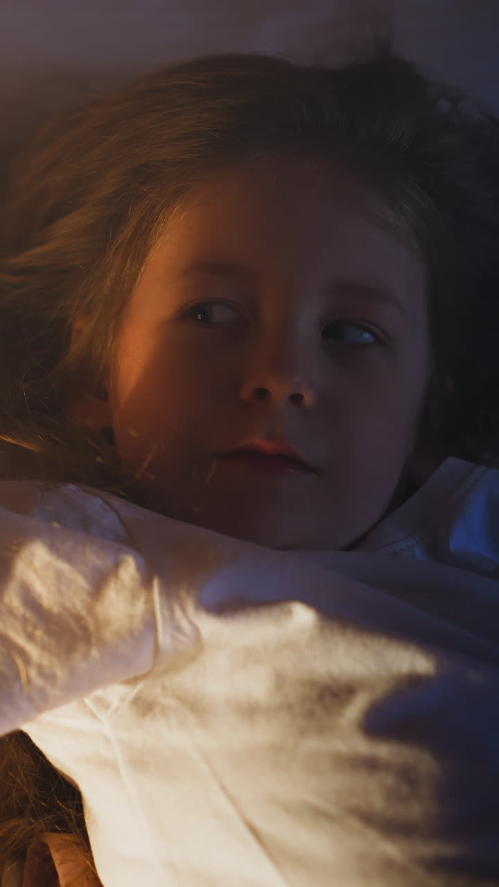 Playful little girl stretches arms on comfortable bed with soft pillows and linen in bedroom partially illuminated by night lamp at home upper view slow motion