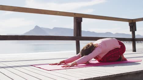 Healthy-mixed-race-woman-practicing-yoga-outdoors,-kneeling-and-stretching-by-the-sea