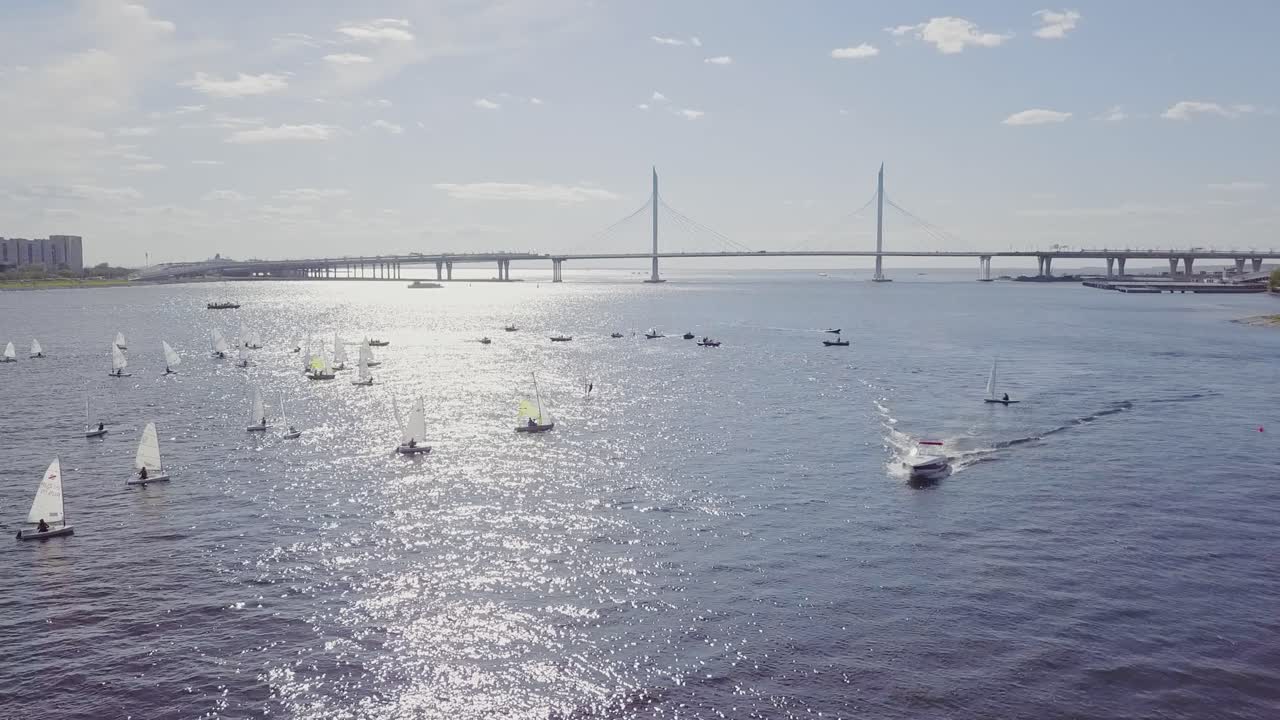 Yachter sejler i en bybugt ved siden af en bro og sommersol, hvide krusninger på vandet.