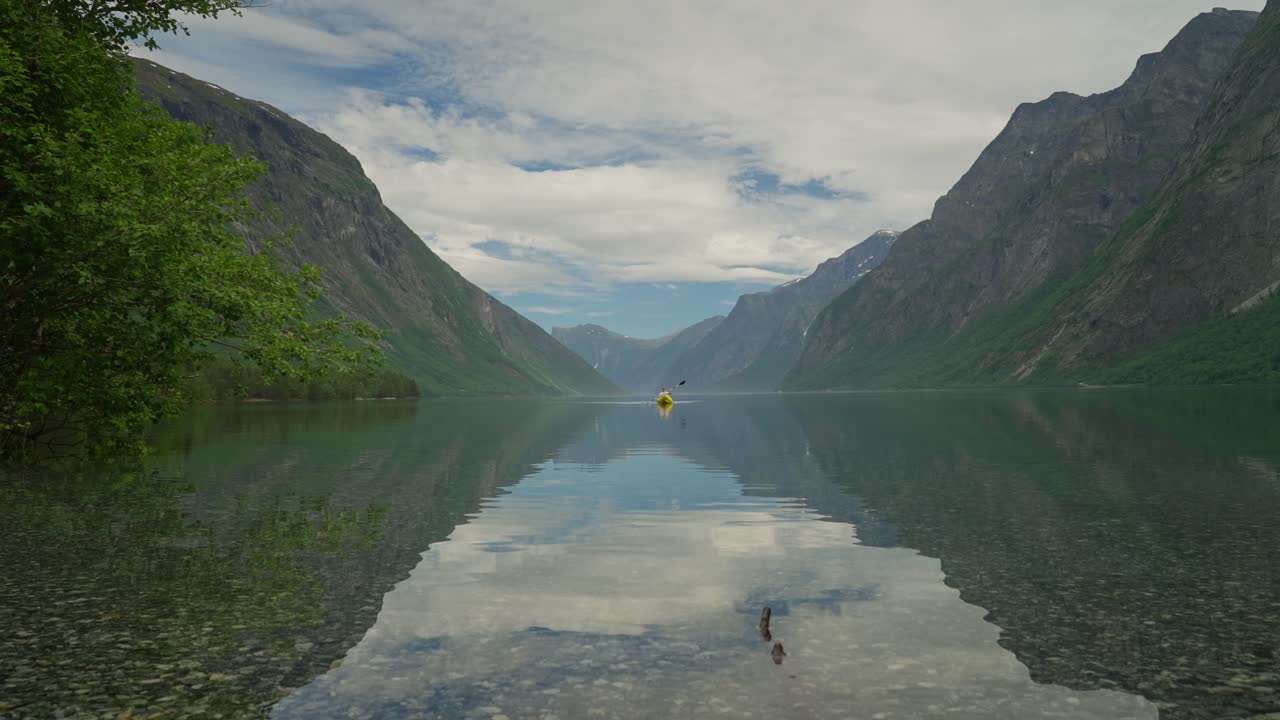 Een gele kajak glijdt in de verte over kristalhelder fjordwater in Noorwegen, Eikesdalsvatnet, dat de omringende bergen weerspiegelt. Serene Noordse scène met kalm water en ongerepte natuur.