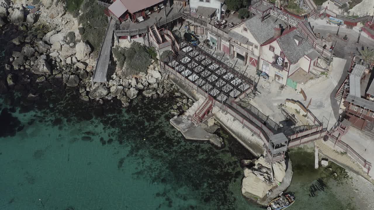 Un drone si allontana da una vista ravvicinata di Popeye Village, sollevando dolcemente la telecamera verso l'alto per rivelare case colorate, Anchor Bay e l'iconica costa di Malta da una prospettiva sempre più ampia.
