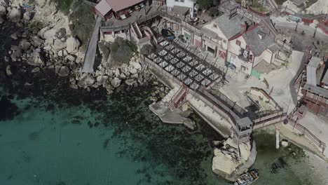 a drone retreats from a close view of popeye village, smoothly lifting the camera upward to reveal colorful houses, anchor bay, and malta’s iconic coastline from an expanding vantage