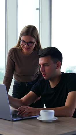 Office teammates cooperating and discussing work. Male and female colleagues looking at laptop talking and smiling. Blurred backdrop. Vertical video