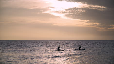 Silhouette-of-Two-Women-Sitting-on-Sup-Boards-Paddleboarding-on-Sea-at-Majestic-Pink-Color-Sunset