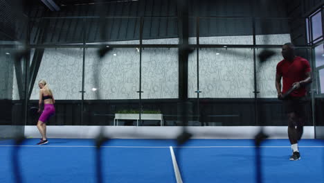 woman playing padel tennis, preparing to hit ball, opponent diving on indoor court