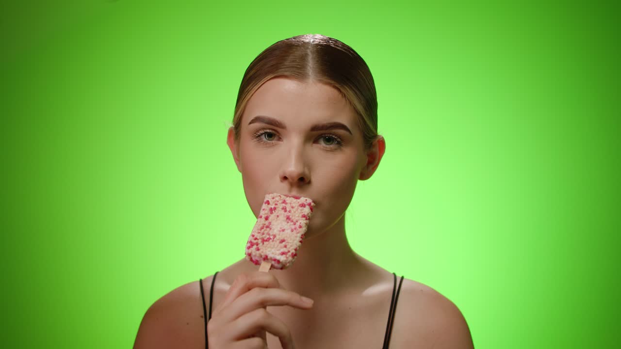 Young caucasian woman takes a bite of a yummy popsicle, smiles, studio shot
