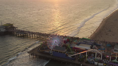 AERIAL:-Coming-down-on-Santa-Monica-Pier-Ferris-Wheel,-Los-Angeles-at-beautiful-Sunset-with-Tourists,-Pedestrians-walking-having-fun-at-theme-park-rollercoaster-with-ocean-view-waves-crashing