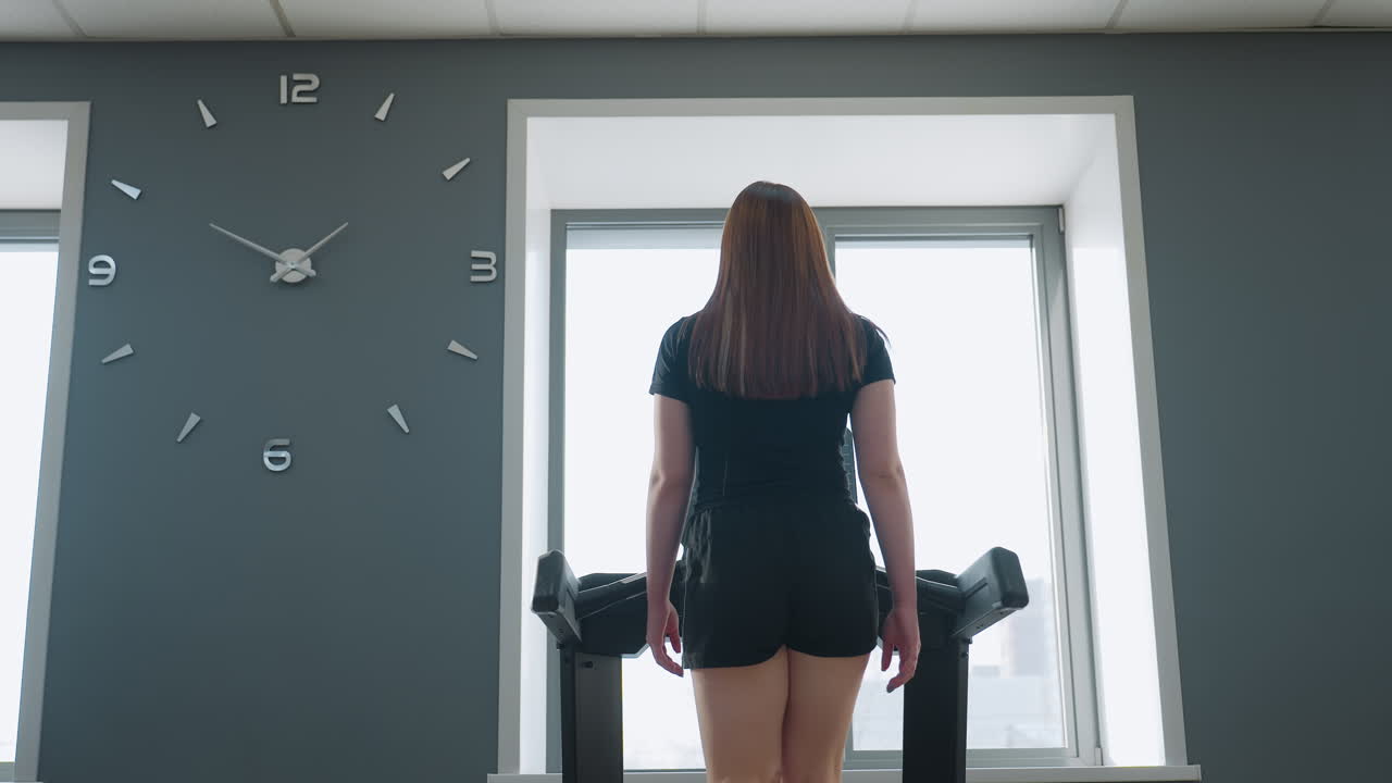woman walking on treadmill in modern gym with residential buildings visible through large window, other treadmill plugged into wall socket on side, soft daylight illuminating clean workout space