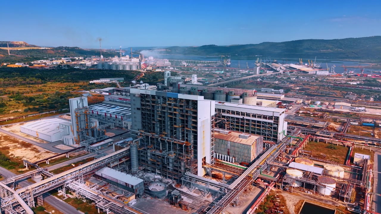 Gran planta de fabricación industrial cerca de un puerto marítimo. Andamios de acero rodean los edificios de la fábrica bajo un cielo azul claro cerca de colinas costeras.