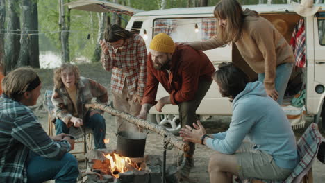 compañía de jóvenes amigos cocinando comida y charlando junto a un fuego de campamento