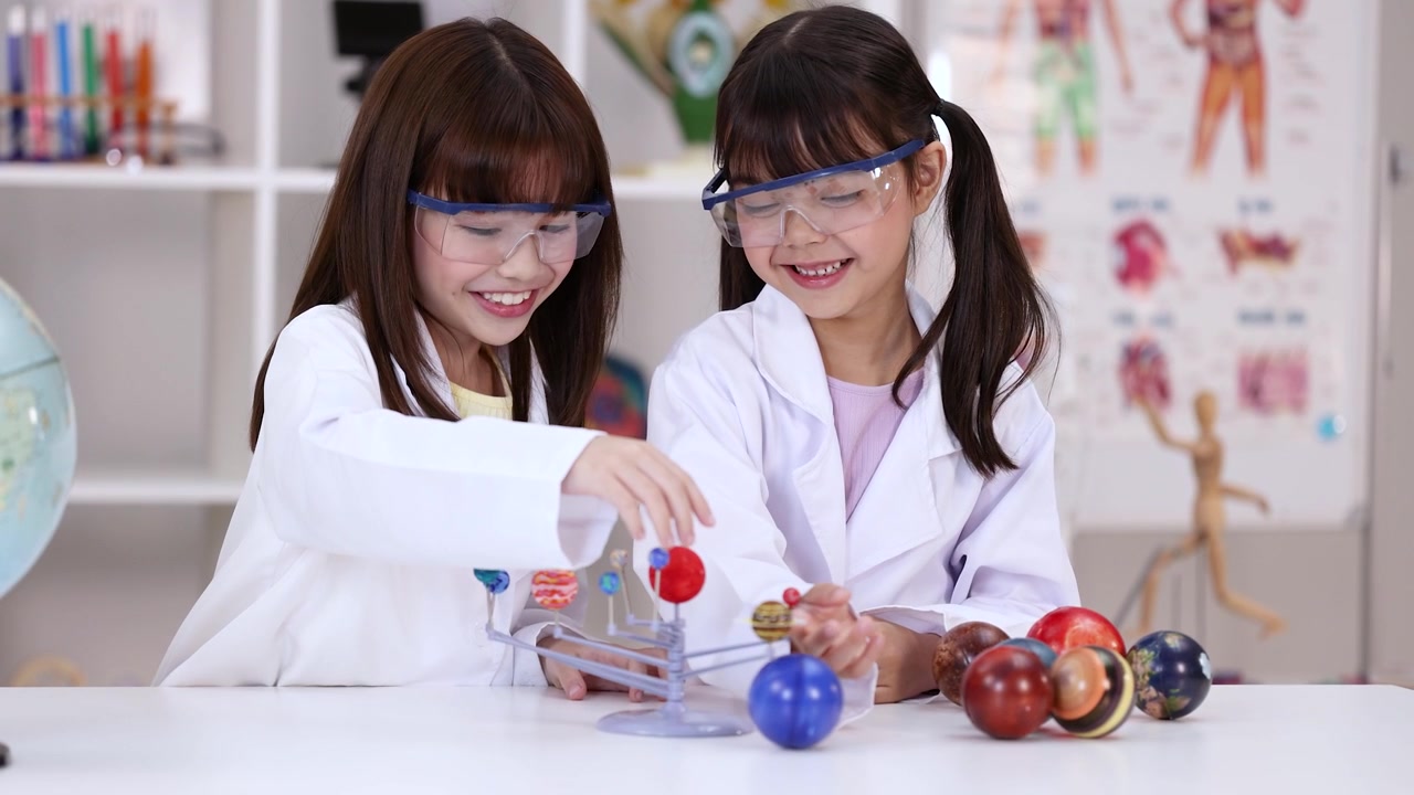 Two smiling girls wearing safety goggles examine a planetary model in a bright science classroom.