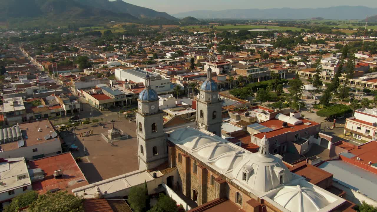 Drone orbit around twin- owered San Juan Bautista temple overlooking town plaza, surrounding rooftops, and distant hills