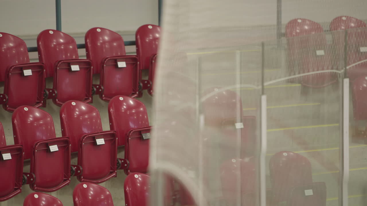 Rack focus from netting above glass in ice rink to empty red folded seats. Establisher shot for sporting event.