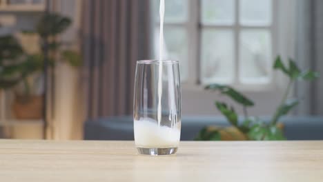 fresh milk being poured in to the glass on the table with home interior in the background
