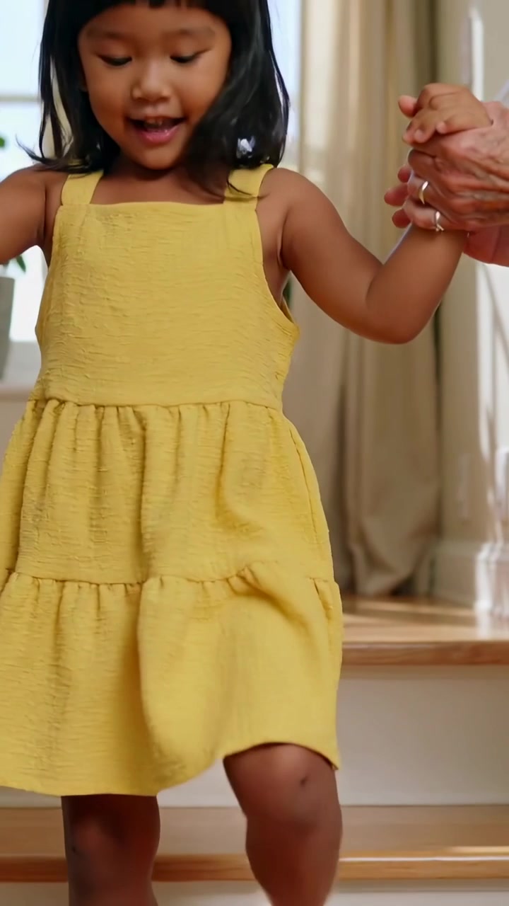 Video verticale: Bambina in abito giallo che scende le scale di casa, mani di un adulto che la guidano.