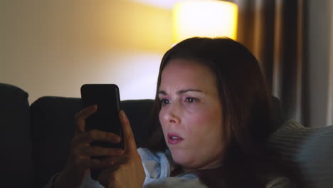 close up of woman lying on sofa at home at night streaming or looking at online content on mobile phone 1