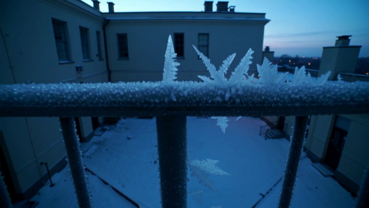 Shifting focus, camera revealing hoarfrost cluster on metal railing in courtyard at dawn, warm lamp