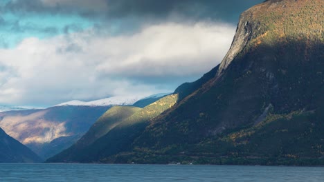 cloudy skies cast soft shadows as sunlight briefly highlights the mountainous slopes above a peaceful fjord