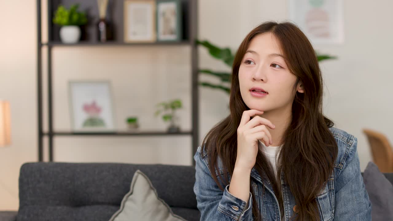 소파에 앉아 턱을 괴고 생각에 잠긴 젊은 여성, 은은한 실내 조명, 고정된 카메라