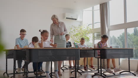 Lehrerin-Kontrolliert-Den-Lernprozess-In-Der-Grundschule.-Eine-Lehrerin-Geht-Zwischen-Den-Schreibtischen-Hindurch-Und-Kümmert-Sich-Um-Die-Erledigung-Der-Aufgabe