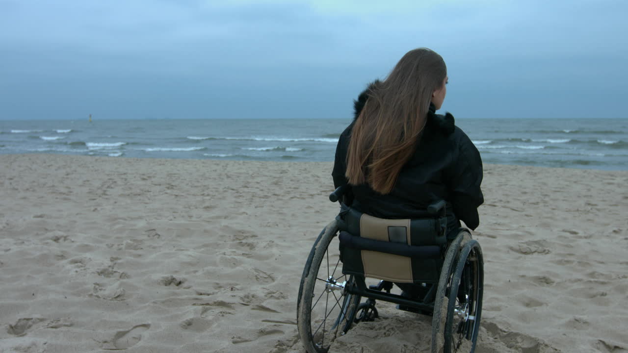 Optimistic disabled woman sitting on wheelchair at the beach on autumn cold day