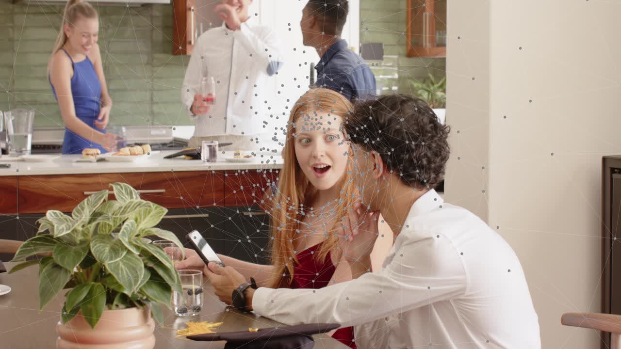 Seated woman reacting to handed phone while man showing shared clip, others serving food, social