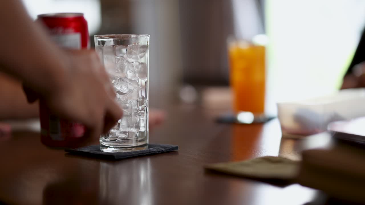 Hand sets red soda can next to ice-filled glass on wooden cafe table, natural lighting