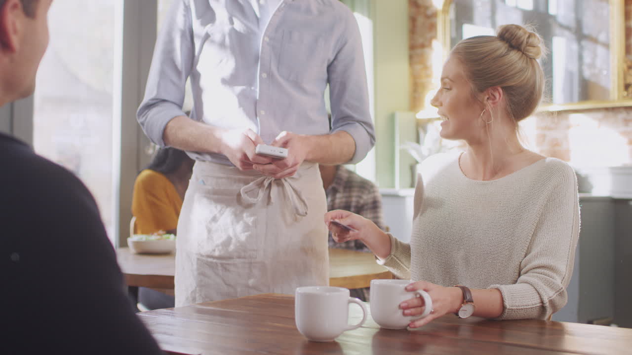 Couple In Coffee Shop Making Contactless Payment With Card For Drinks To Male Waiter