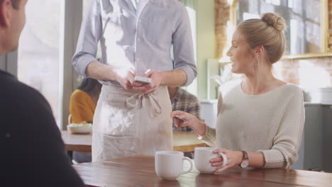 Couple-In-Coffee-Shop-Making-Contactless-Payment-With-Card-For-Drinks-To-Male-Waiter