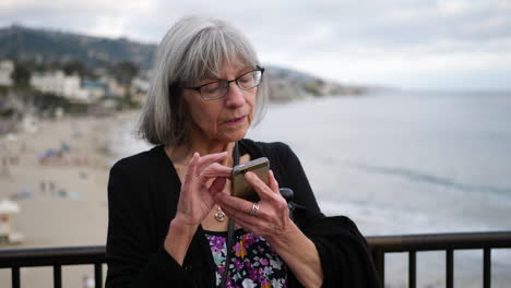 a middle aged woman with aging white hair and eye glasses texting on her phone on vacation in laguna beach, california slow motion