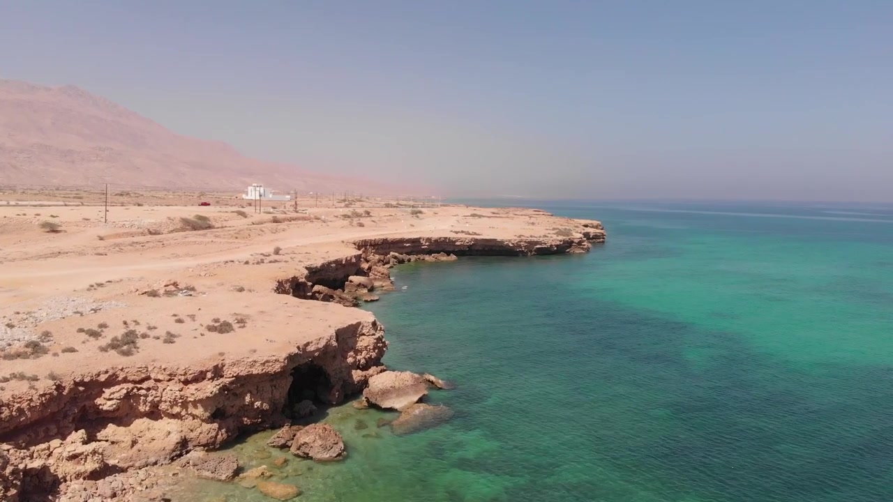 Ripresa aerea con drone dall'alto di una strada costiera tortuosa lungo le onde turchesi dell'oceano e le aspre scogliere desertiche dell'Oman. Mette in mostra il drammatico contrasto tra paesaggio arido e mare.