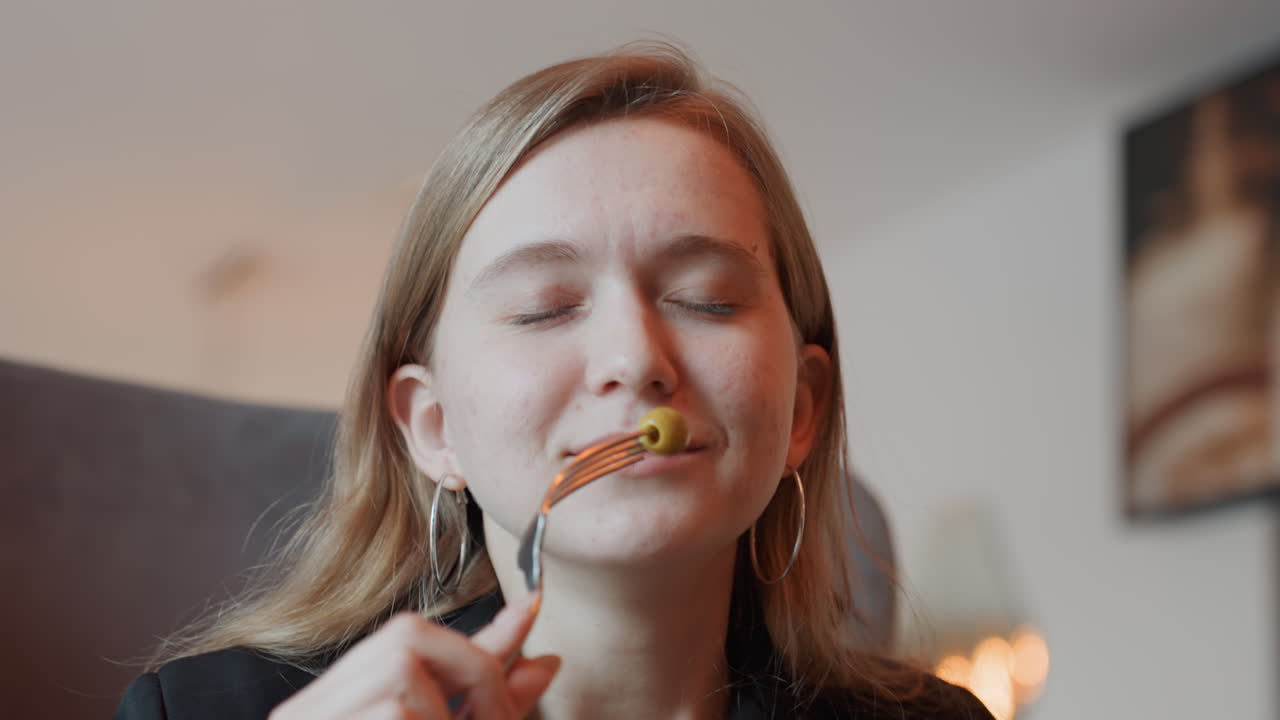 Joven mujer caucásica probando una aceituna, sonriendo con los ojos cerrados, sosteniendo un tenedor, en una sala de estar acogedora con luz cálida de lámpara, ambiente de bloguera de comida, degustación lúdica de un deleite sutil, saboreando lentamente, un momento de estilo de vida cercano