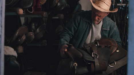 old man inspecting saddles in a dusty stable