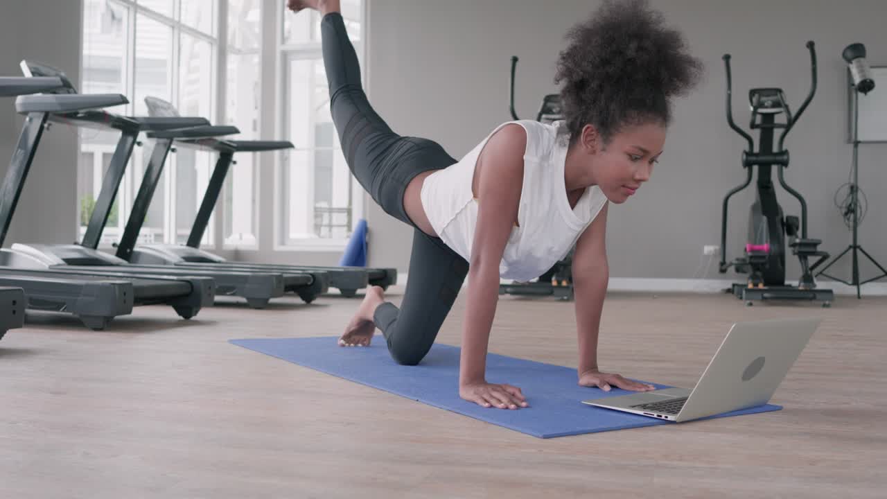 fitness joven mujer negra haciendo ejercicio yoga en el gimnasio. mujer afroamericana loook en la lección de computadora portátil practicando la postura de tabla yaga en la clase de internet en línea social. concepto de estilo de vida saludable.