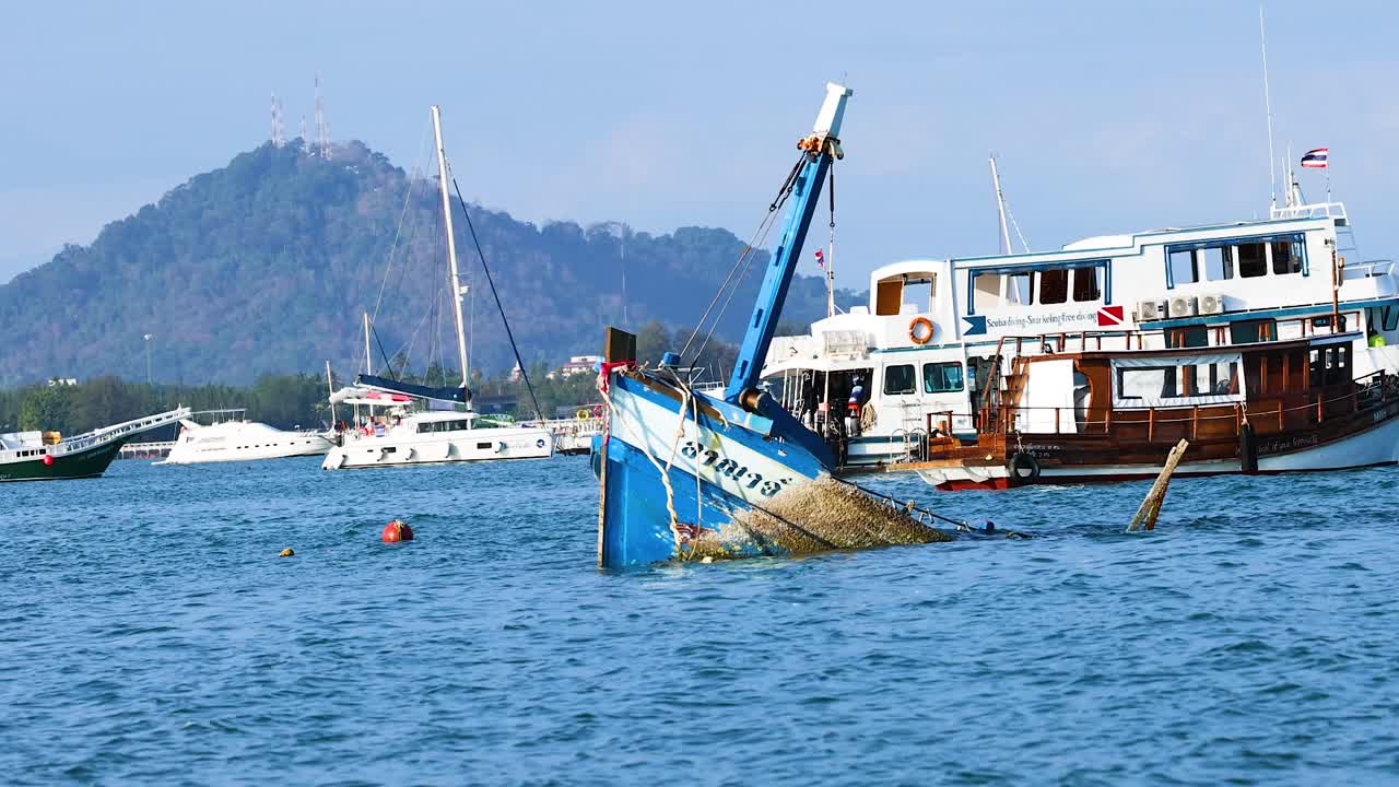 A fishing boat partially submerged in Phuket harbor, surrounded by other vessels under clear skies