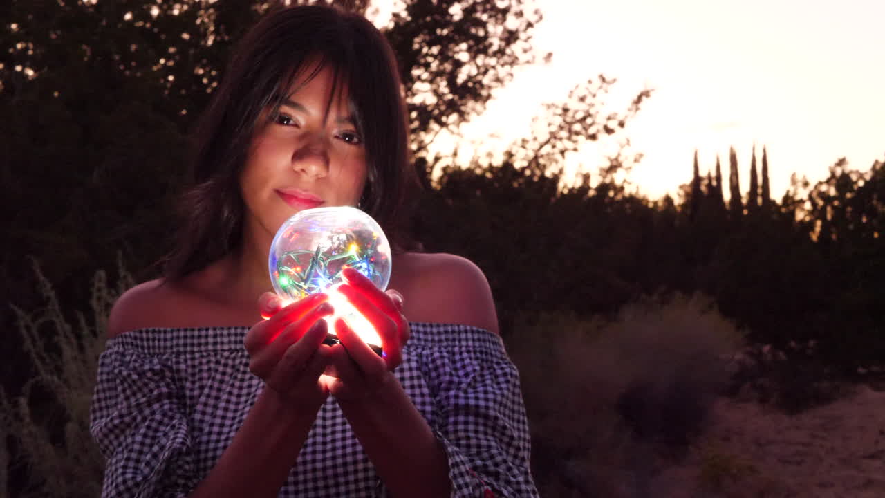 A beautiful woman with a magic glowing crystal ball illuminated by lights in the woods at sunset.