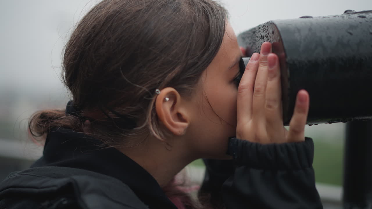 Close-up portret toont vrouw die tuurt, druppels volgen wang en haarlijn, vingers omlijsten oculair, ogen gericht op verre skyline, zacht licht onder bewolkte hemel, emotionele mix van verwondering en nostalgie.