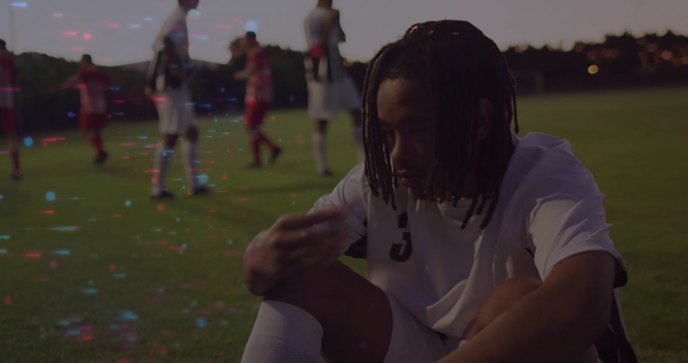 Soccer player reacting to failed play by sitting dejected on grass at dusk with teammates looming