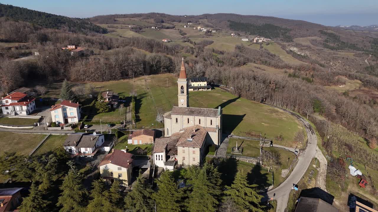 Il campanile dei Santi Salvatore e Gallo si erge sopra la tranquilla I Rabbini, il villaggio di Morfasso, la tradizionale chiesa in pietra e le case immerse nelle verdi valli appenniniche sotto un cielo sereno e soleggiato