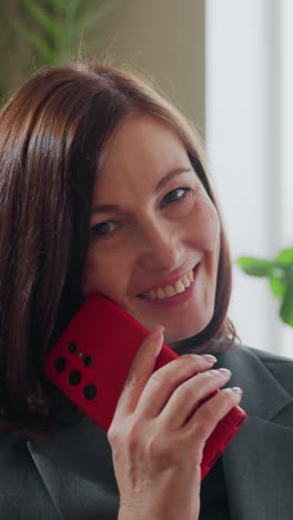 Smiling businesswoman in gray blazer sitting on couch holding red smartphone looking confidently at camera in bright modern office space with green indoor plants and natural window lighting