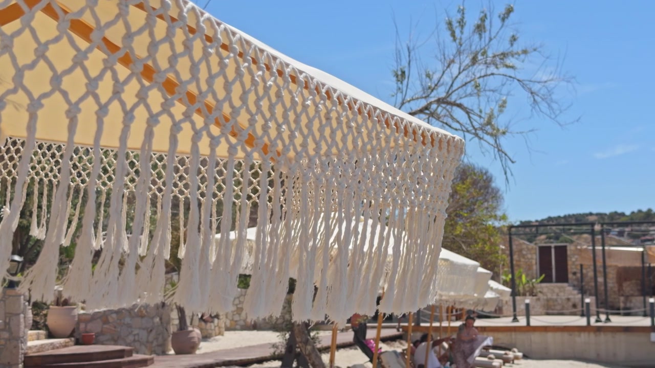 Close-up shot of a fringed macrame umbrella and white cabana drapes on a wooden beach deck, overlooking a turquoise bay and distant green hills.