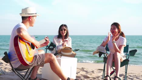 happy young people enjoying with music at the beach, young man playing guitar and singing as his friends clapping and laughing, having fun on the beach picnic in summer holidays, vacation.