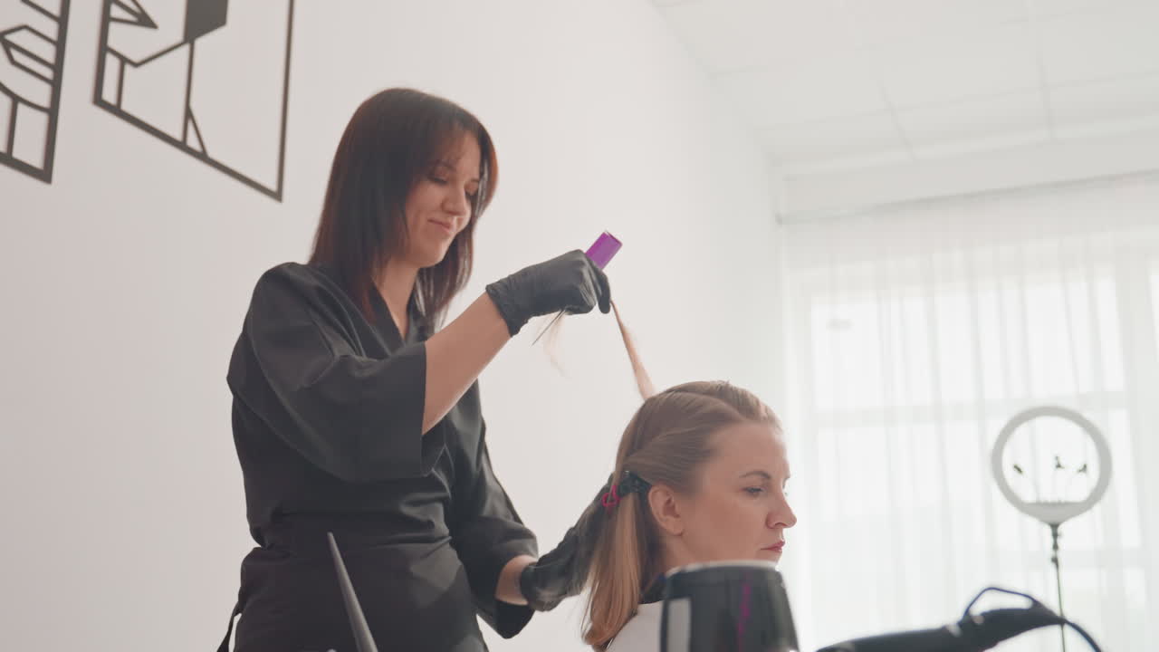 Estilista aplicando tinte para el cabello en un estudio luminoso, clienta sentada en una silla, estilista con guantes negros separando el cabello y aplicando el color con un pincel y papel de aluminio, luz natural suave a través de una tela fina