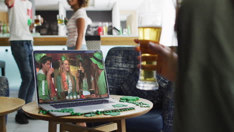 person at bar using laptop making st patrick's day video call with friends at another bar