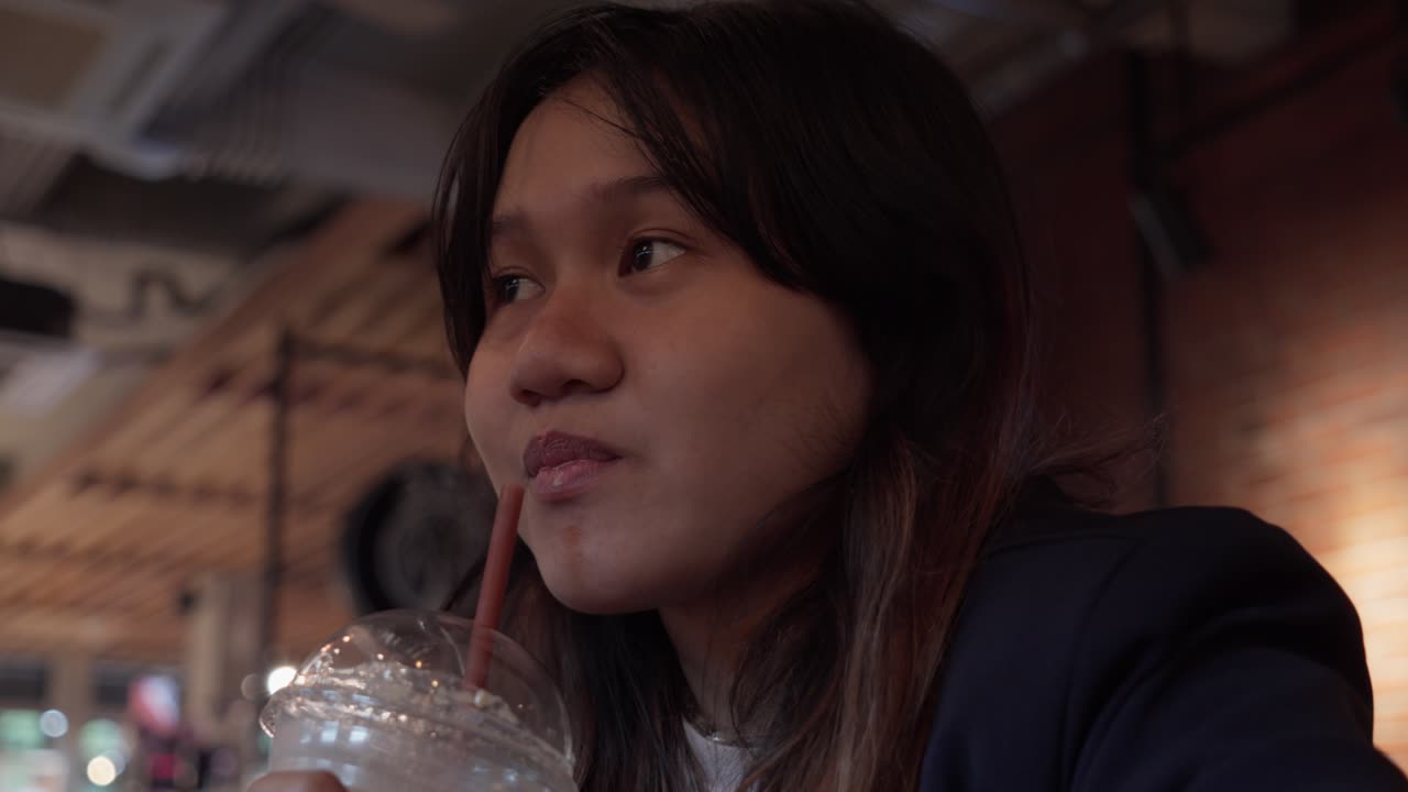 A young woman sipping a cold drink, gazing thoughtfully into the distance, indoors