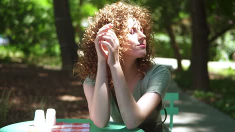 Retrato-De-Una-Sensual-Chica-Pelirroja-Rizada-Tocando-Su-Cabello-Rojo-En-Verano-Mientras-Está-Sentada-En-Un-Parque-Al-Aire-Libre-Con-Destellos-De-Lente.-Disfrutando-Del-Tiempo-Al-Aire-Libre