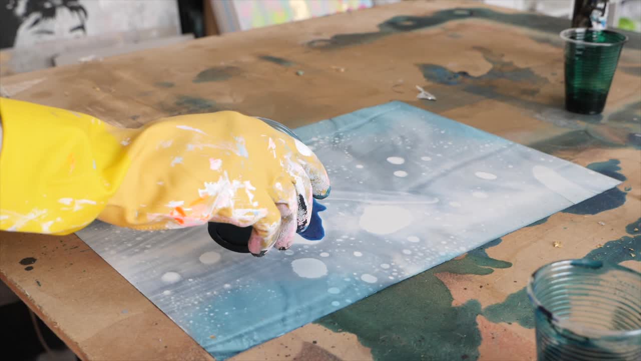 Artist hand with yellow glove pours blue tint on sheet surface. Slow-motion