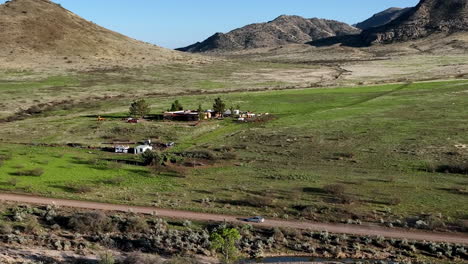 Aerial-shot-of-car-driving-on-dirt-road-in-Willcox,-Arizona,-wide-side-angle-drone-shot-with-home-and-mountains-in-the-background