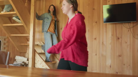 three girls walking down wooden staircase inside rustic cabin interior, paper shopping bags on wooden table in foreground, cozy sunlight casting shadows on timber wall